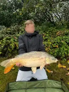 A boy crouching down at the edge of a lake holding a large fish that he has caught