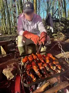 A boy sat cooking chicken drumsticks over an open fire in the woods