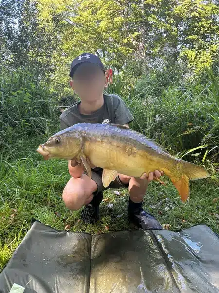 A boy crouching down at the edge of a lake holding a large fish that he has caught