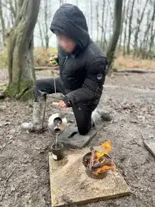 a boy pouring a hot pot of water into a mug in front of an open fire in the woods