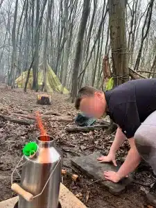 A boy crouching down next to a metal flask in the woods
