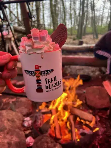 A mug of hot chocolate with cream and marshmallows being held in front of an open fire in the woods