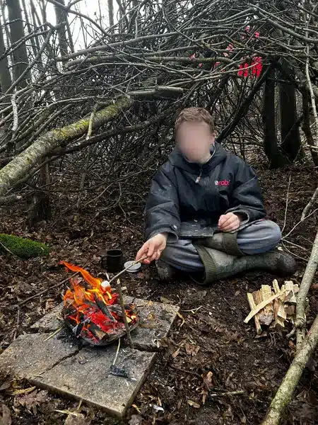 A boy sitting at the entrance to a makeshift shelter holding a marshmallow on a stick over an open fire