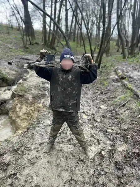 A boy standing in deep mud in the woods in just his socks holding his muddy shoes