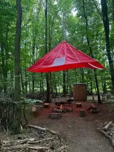 A seating area in the woods with small logs as seats and a large red tarpaulin suspended above