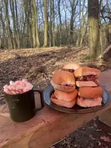 A mug of hot chocolate with marshmallows poking out the top next to a stack of bacon rolls on a bench in the woods