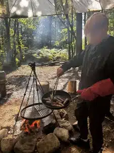 A boy cooking sausages in a pan over an open fire in the woods