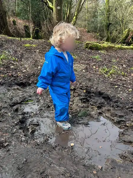 A small boy wearing waterproofs walks through a muddy puddle in the woods