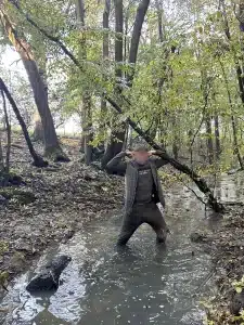 A boy stood in a stream in the woods putting his hands up to the side of his head