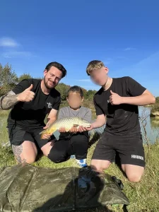 A boy crouching down at the edge of a lake holding a large fish that he has caught, flanked by two others