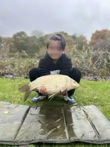 A boy crouching down at the edge of a lake holding a large fish that he has caught