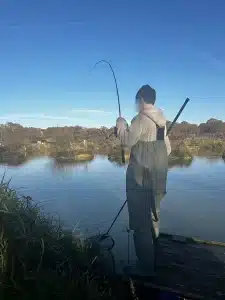 A boy fishing on a small jetty on the edge of a lake holding a rod which is bending