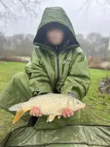 A boy crouching down at the edge of a lake holding a large fish that he has caught
