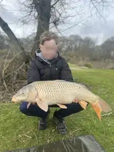 A boy crouching down at the edge of a lake holding a large fish that he has caught