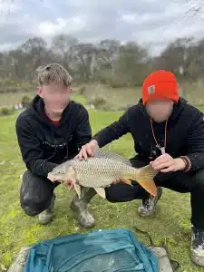Two boys crouching down at the edge of a lake holding a large fish that they have caught