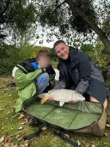 A boy and Ben, a Trail Blazers staff member, crouching down and displaying a fish they have caught from a lake