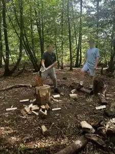 A boy chopping wood with an axe in the woods as another boy looks on