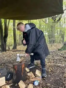 A boy chopping wood with an axe as he gives a thumbs up to the camera